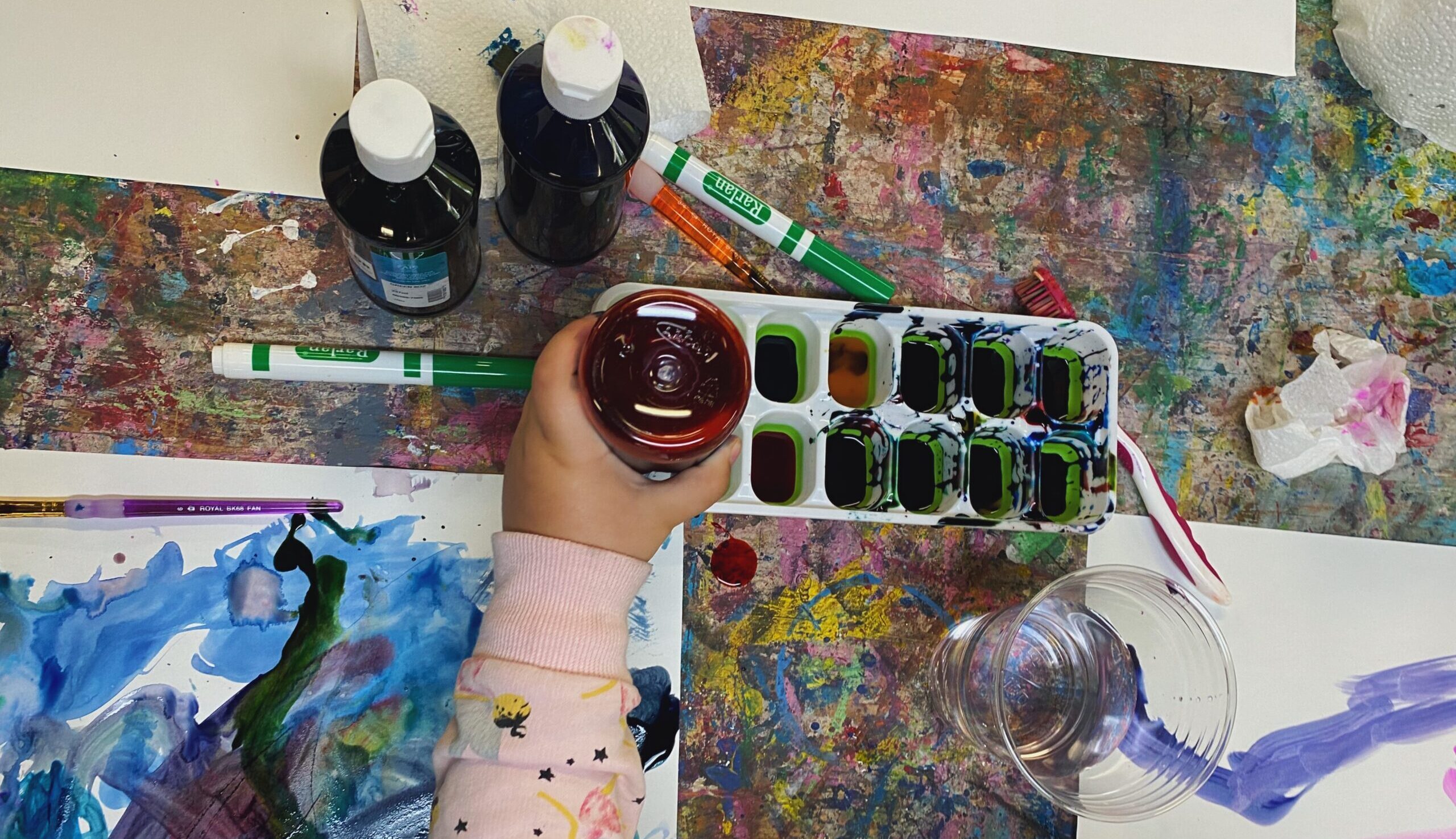 Umbrella Healing Together, Vermont Network Against Domestic & Sexual Violence (DV/SV Coalition) photo of a child hand and arm holding a bottle of paint on a colorfully painted table.