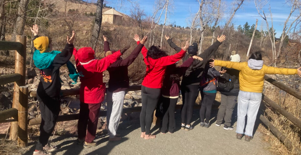 A group of nine people with their backs to the camera lift their arms out to the sky.